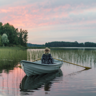 Une femme coiffée d’un chapeau est assise dans un canot à rames tandis qu’un lac et des étendues forestières sont visibles à l'arrière-plan.