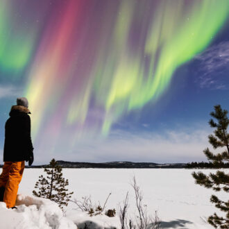 Una persona ataviada con ropa de invierno se ha detenido sobre unas rocas cubiertas de nieve para contemplar una aurora boreal verde y púrpura que se expande por el cielo.