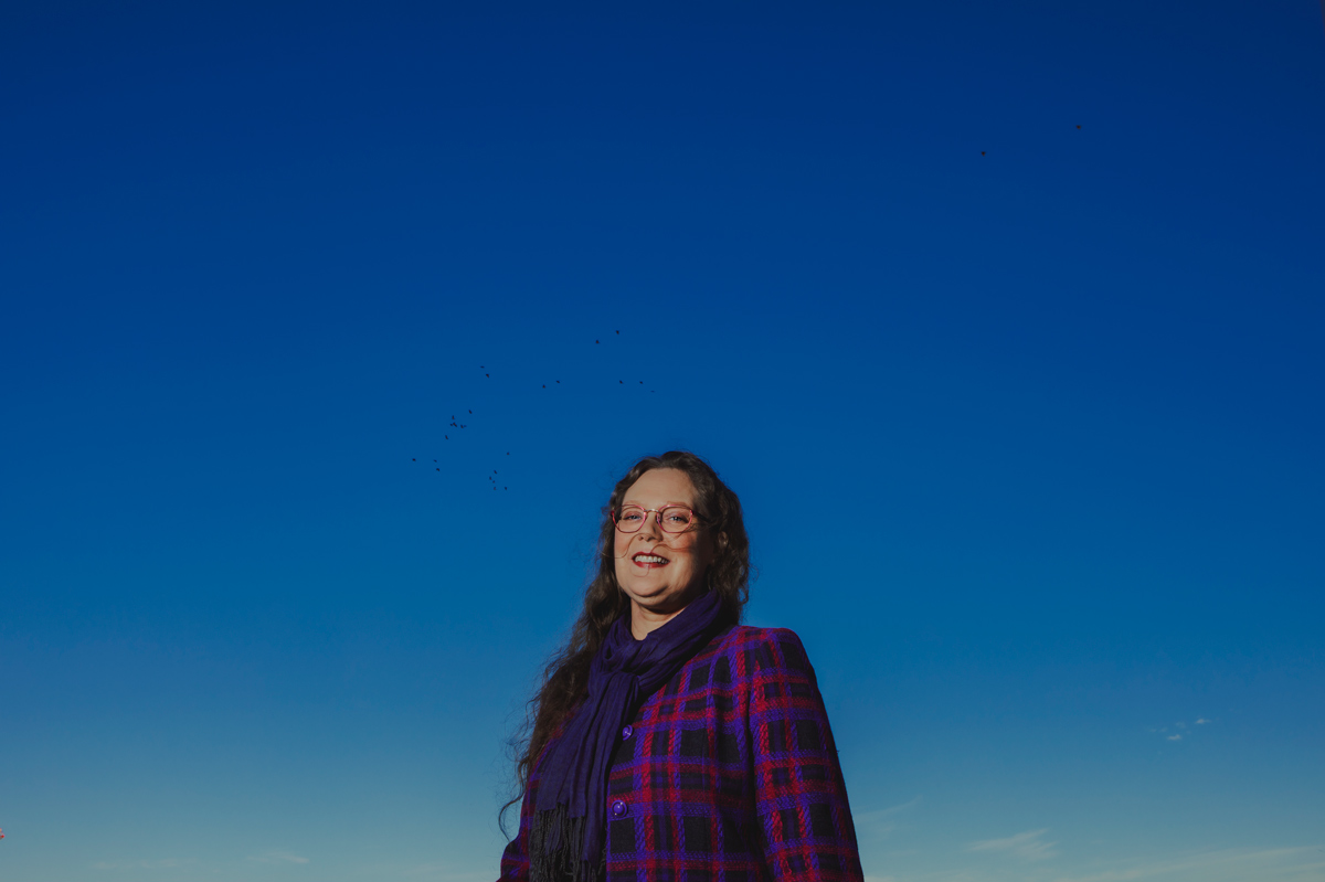 A woman wearing a jacket and scarf stands beneath a blue sky.