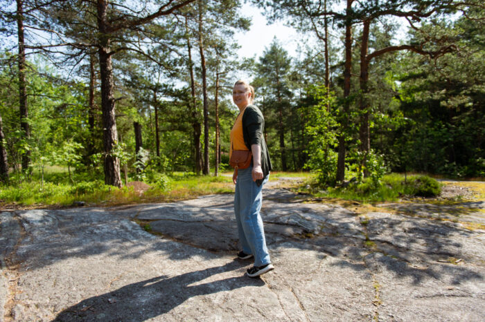 A woman walks across a sunlit rock in a clearing beneath tall pines.