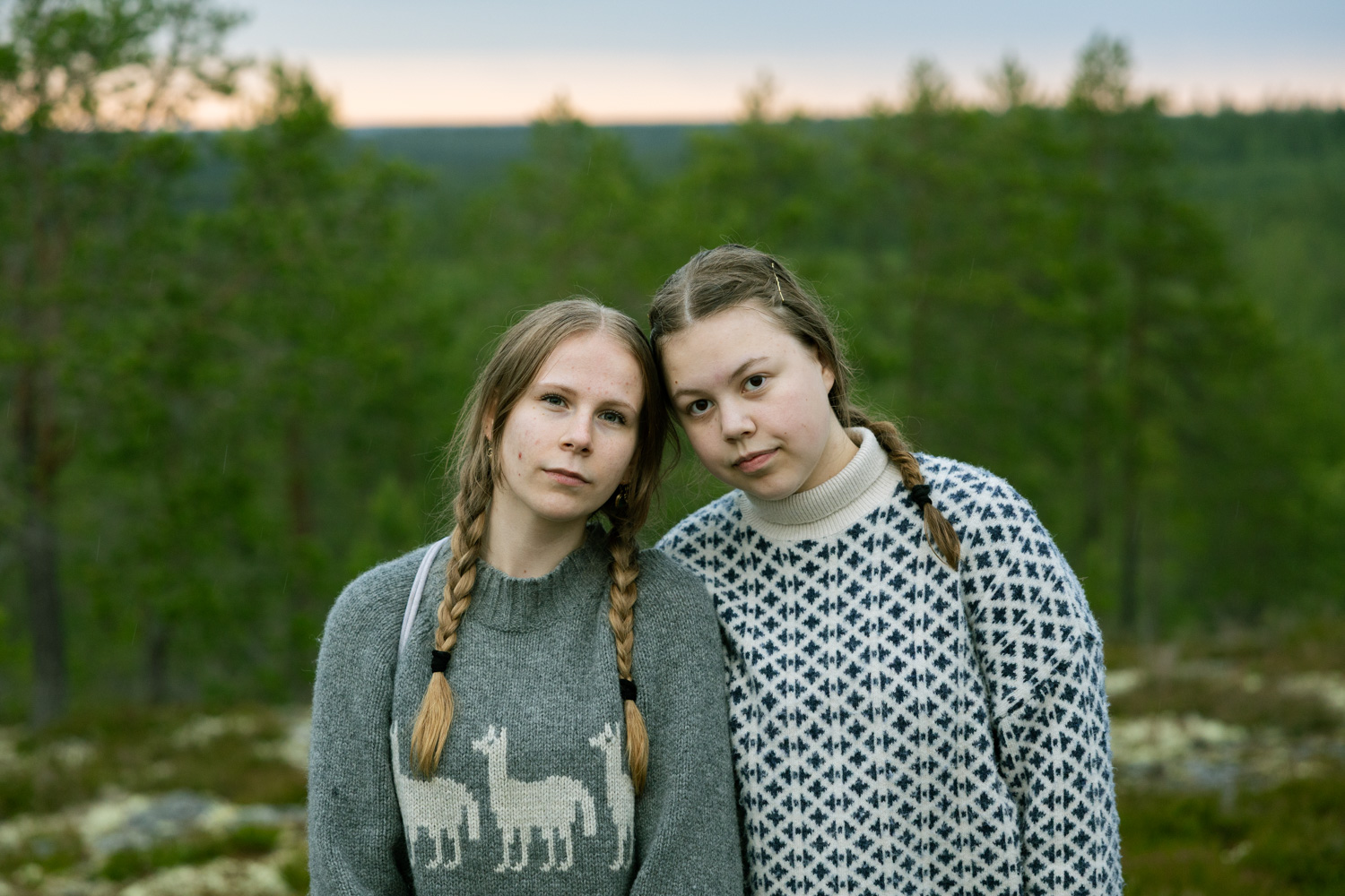 Two young women pose with heads together in front of a forested landscape falling away into the distance.