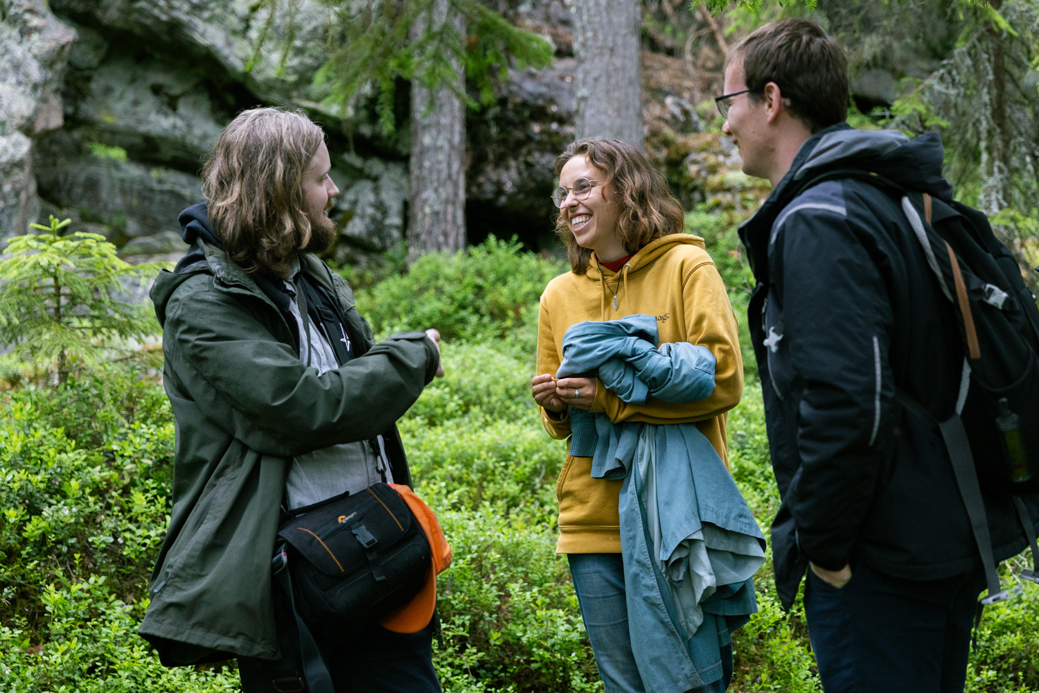 Three smiling people talk in front of a forested cliff.
