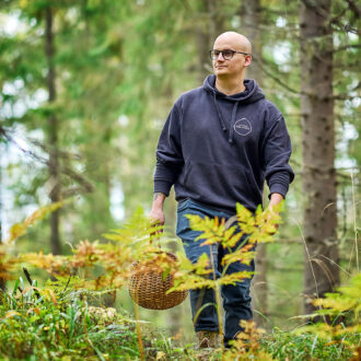 Um homem caminha por uma floresta, fora da trilha, carregando uma cesta de vime para colher alimentos. Samambaias e plantas silvestres crescem densamente ao seu redor, sob árvores altas.