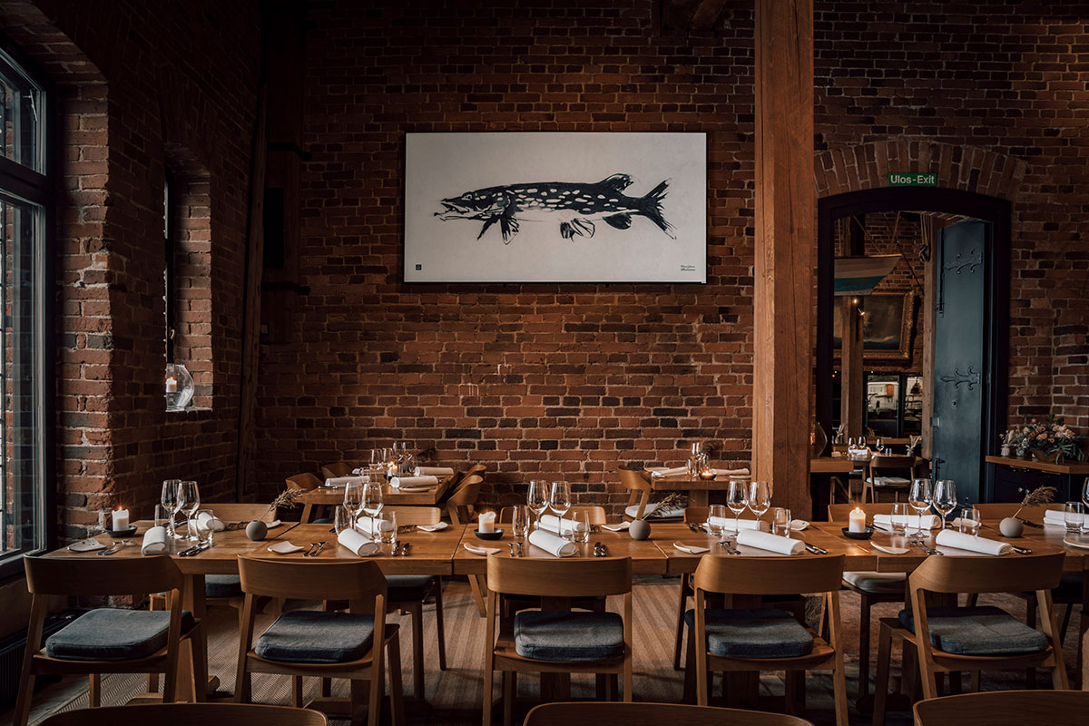 Interior de um restaurante com paredes de tijolos, mesas de madeira e uma pintura representando um peixe da espécie lúcio na parede ao fundo.