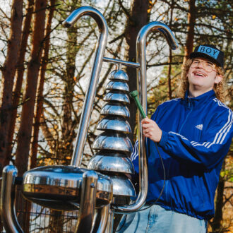 Un homme fait mine de jouer d’un grand instrument de musique en acier installé sur une aire de jeux extérieure.