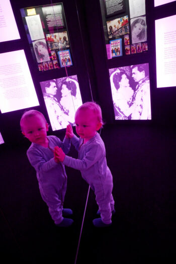 A small child leans curiously against a mirror at a museum.