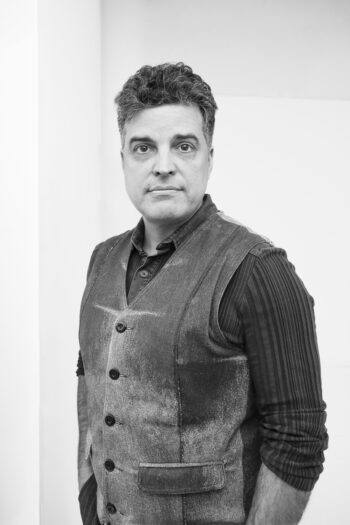 Black and white portrait of a man, hands in pockets, looking toward the camera.