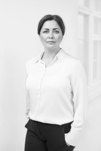 In a black and white portrait, a woman looks at the camera with her hands in her pockets.