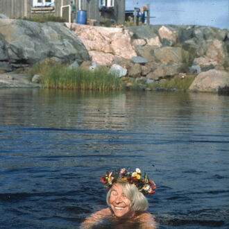 La tête ceinte d’une couronne de fleurs, une femme est en train de nager dans la mer, tandis qu’un rivage rocheux et un petit chalet d’été sont visibles derrière elle.