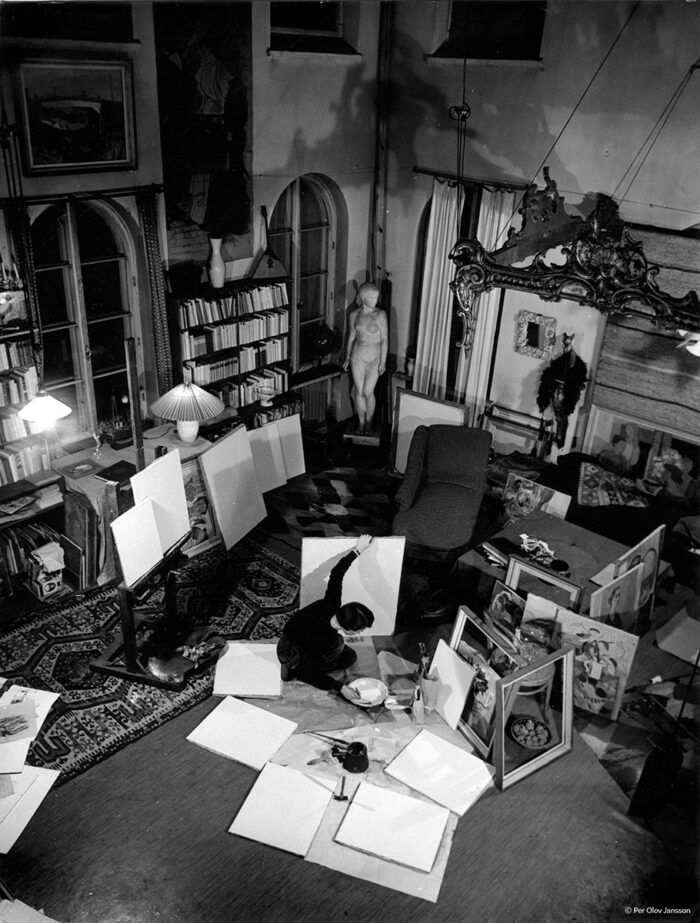 A woman kneels in an art studio, surrounded by blank canvases. She dips a brush into paint on the floor while holding a canvas steady with her other hand.