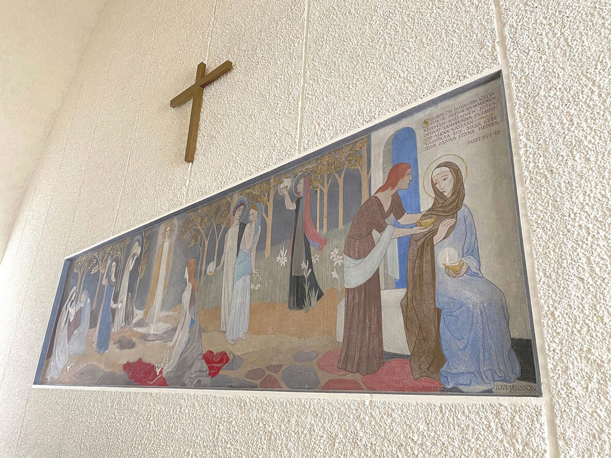 A colourful mural shows ten women holding lanterns, painted on a white wall above an altar and beneath a metal cross.