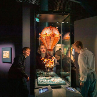 Visitantes de um museu examinam um modelo de balão de ar quente listrado suspenso com pequenos sacos de areia dentro de uma vitrine de vidro.