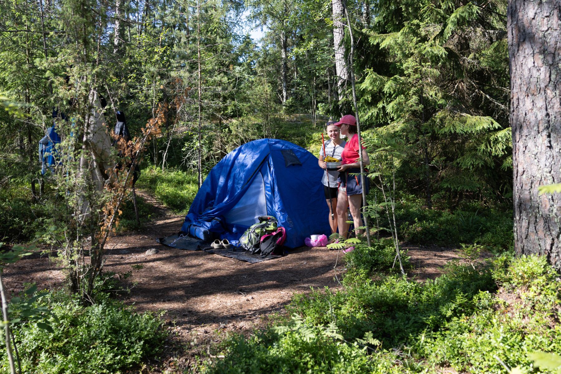 Two women stand beside a blue dome tent pitched in a lush green forest.