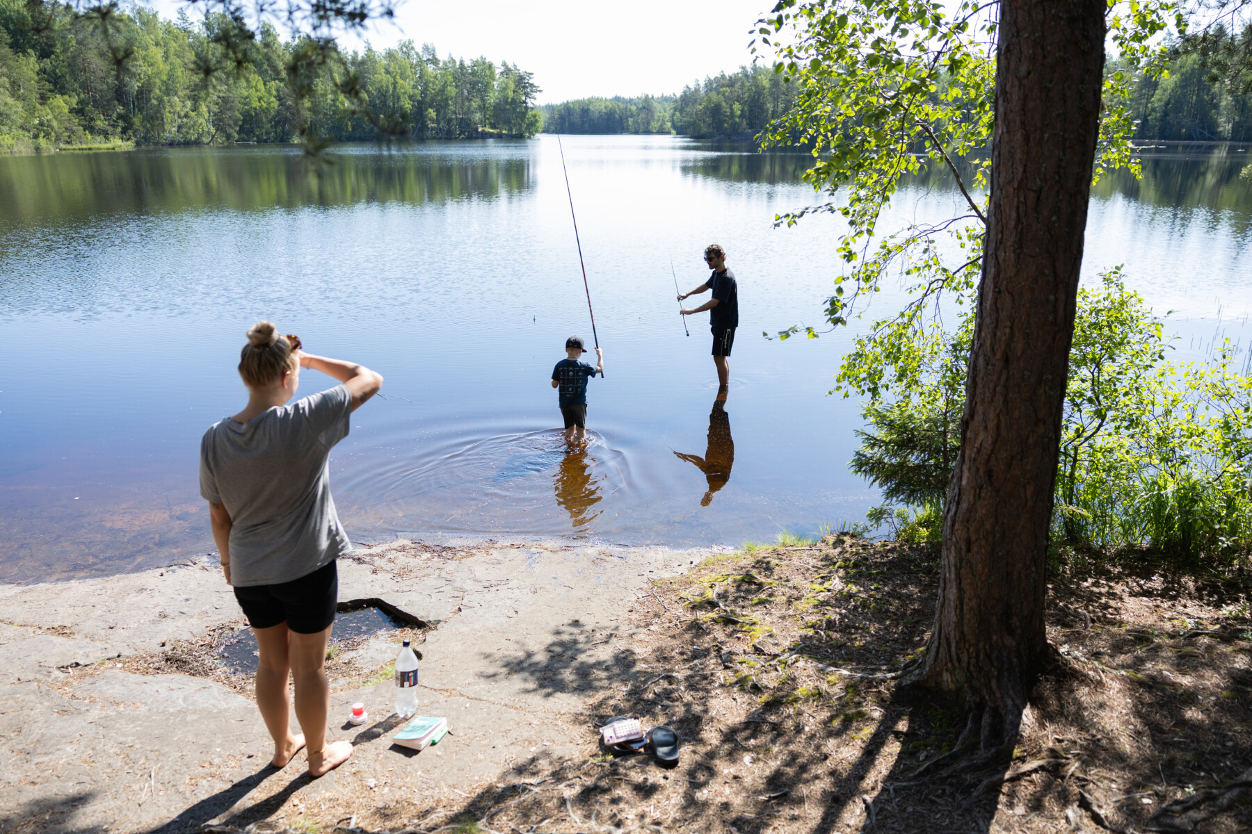 A man and a child stand in shallow lake water holding fishing rods while a woman watches from the shore.