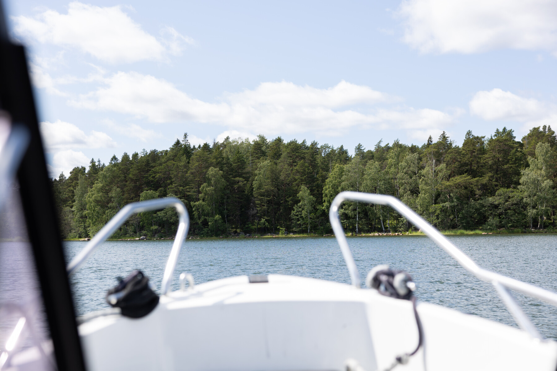 The front of a small white boat with railings approaches a forested shoreline across calm water.