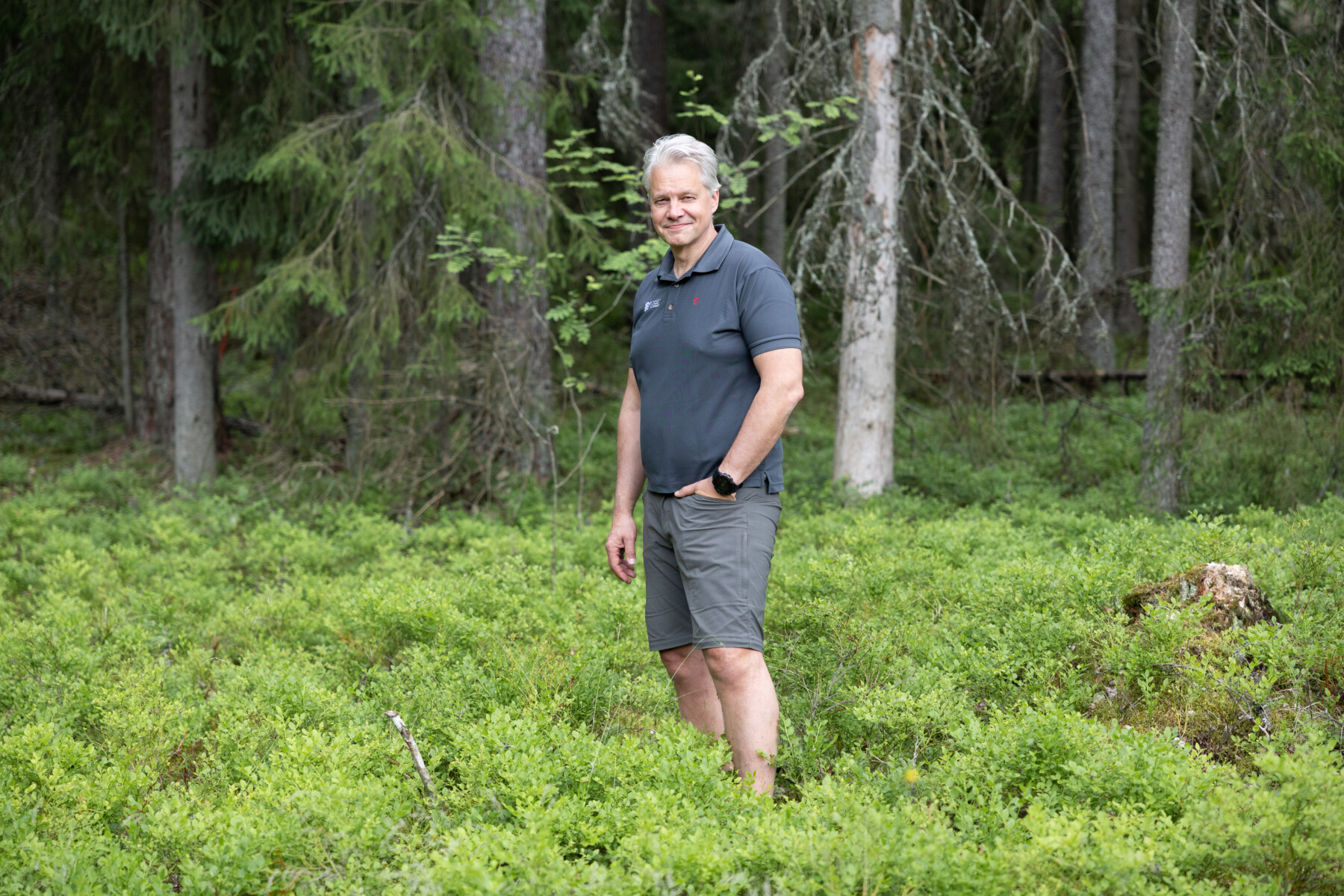 A man stands in a leafy green forest surrounded by trees and low vegetation.