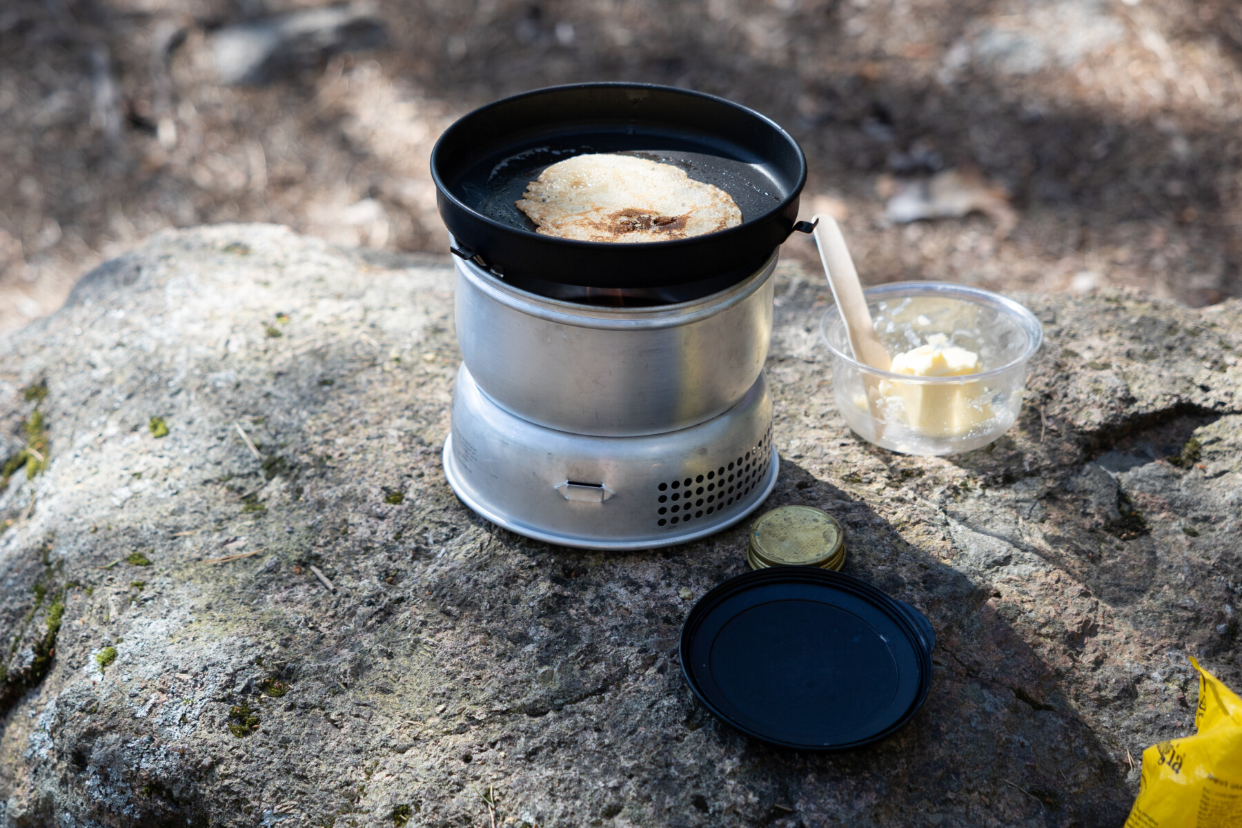 A camping stove rests on a flat rock with a frying pan heating food in a forest setting.