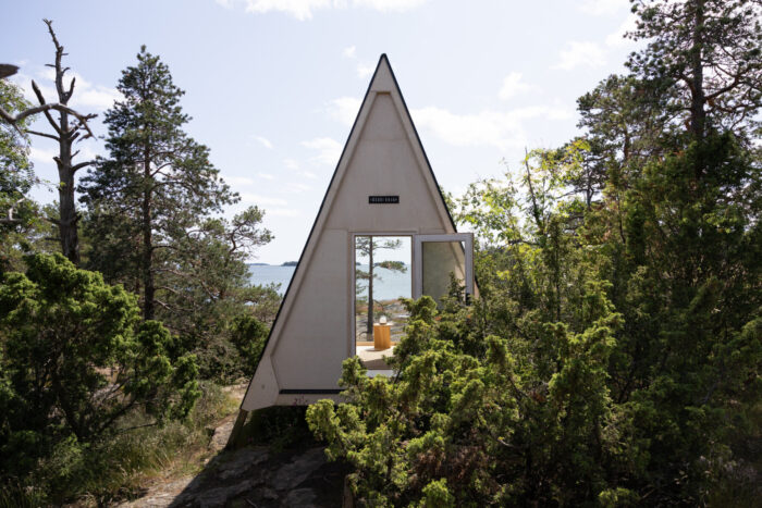 A triangular wooden cabin sits among bushes with its door open and a view of the sea through trees.