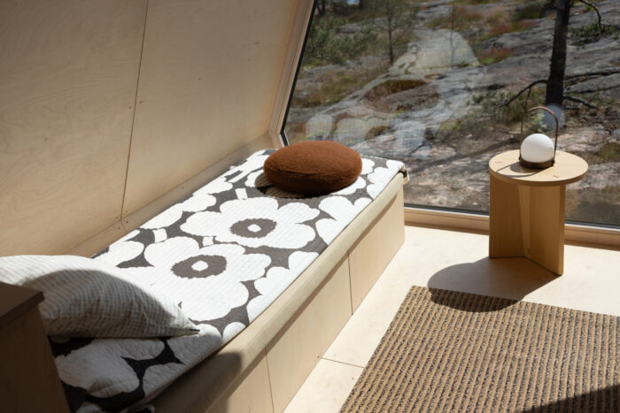 A simple cabin room features a bed with floral bedding, wood-paneled walls and floors and a large window onto the forest outside.