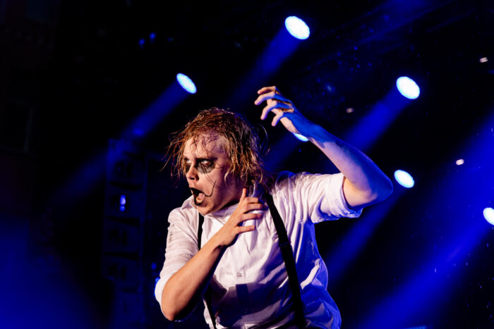 A man with dark eye makeup holds up an imaginary guitar under bright lights.