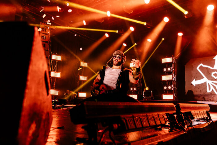 A man in a fur hat and sunglasses slides on his knees on stage as he plays an imaginary guitar.