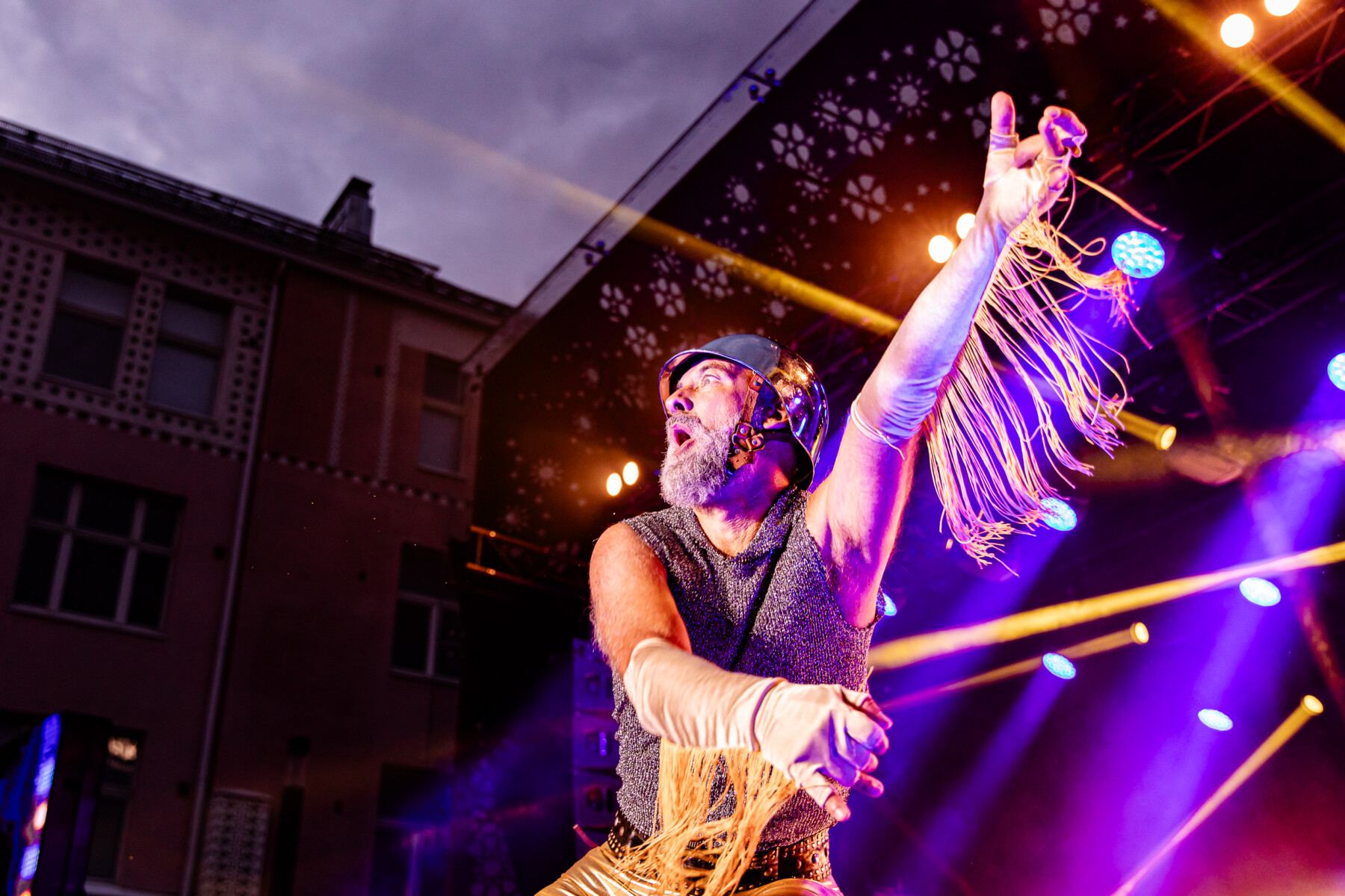 A bearded man wears a helmet and tasseled gloves as he plays an imaginary guitar.