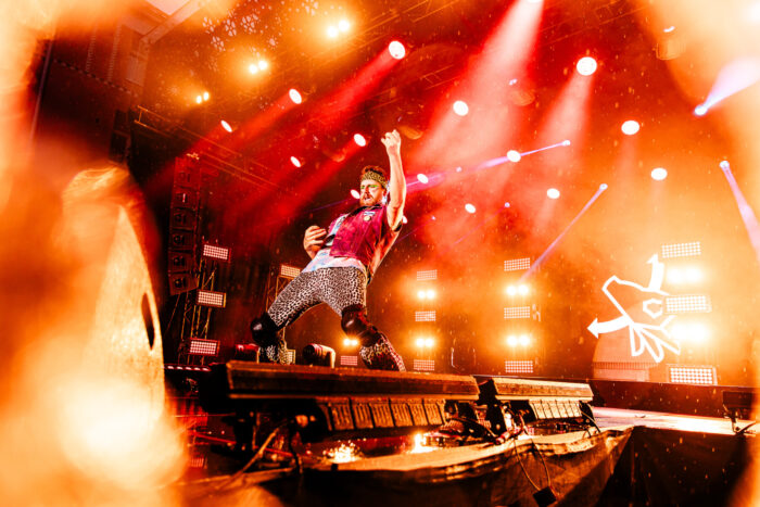 A man in a red shirt, spotted pants and knee pads plays an imaginary guitar on a stage.