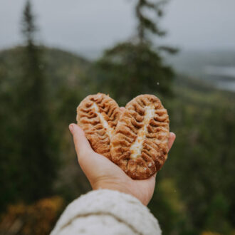 An outstretched hand holds two small oval-shaped pies in front of a background of trees and a lake.