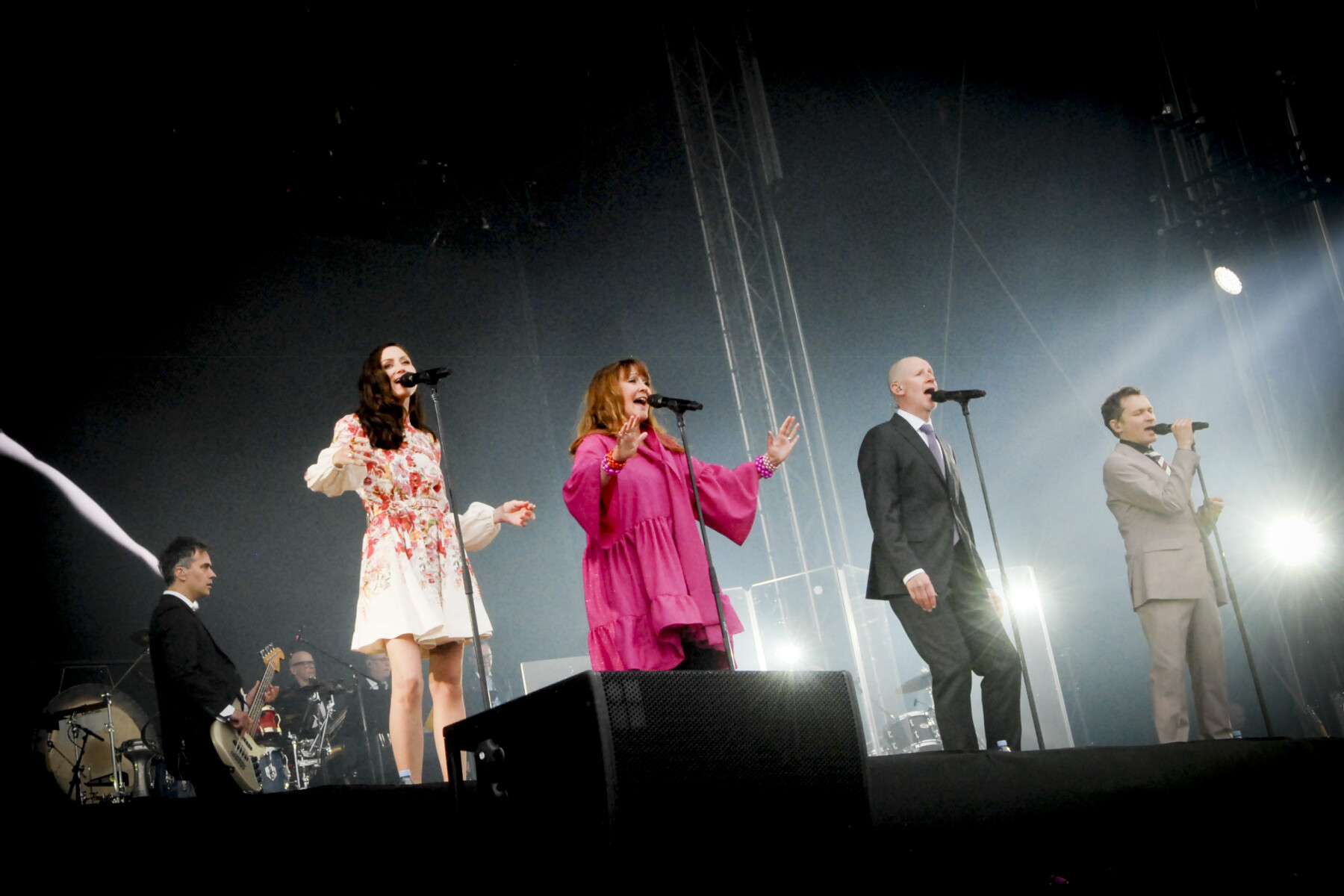 Two women in dresses and two men in suits stand onstage singing with a bassist and drummer in the background.