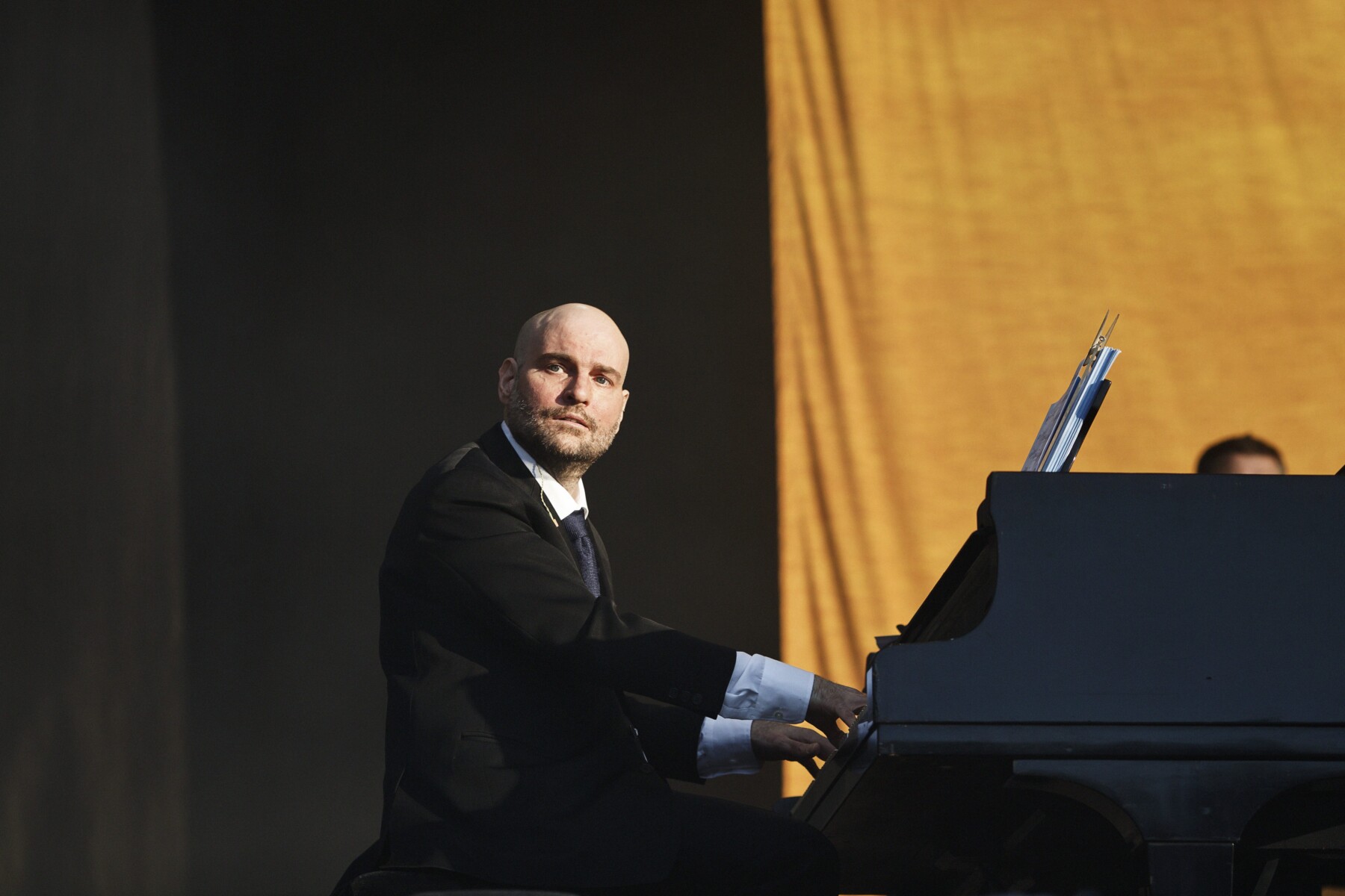 A man in a black suit looks toward the camera while playing a grand piano onstage.