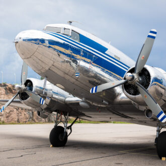 Um avião DC-3 vintage com listras azuis e brancas pousa em um campo de pouso sob um céu nublado.