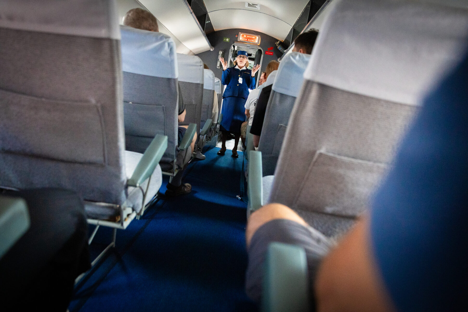 Uma comissária de bordo está no corredor da cabine de um avião antigo, dando instruções aos passageiros.