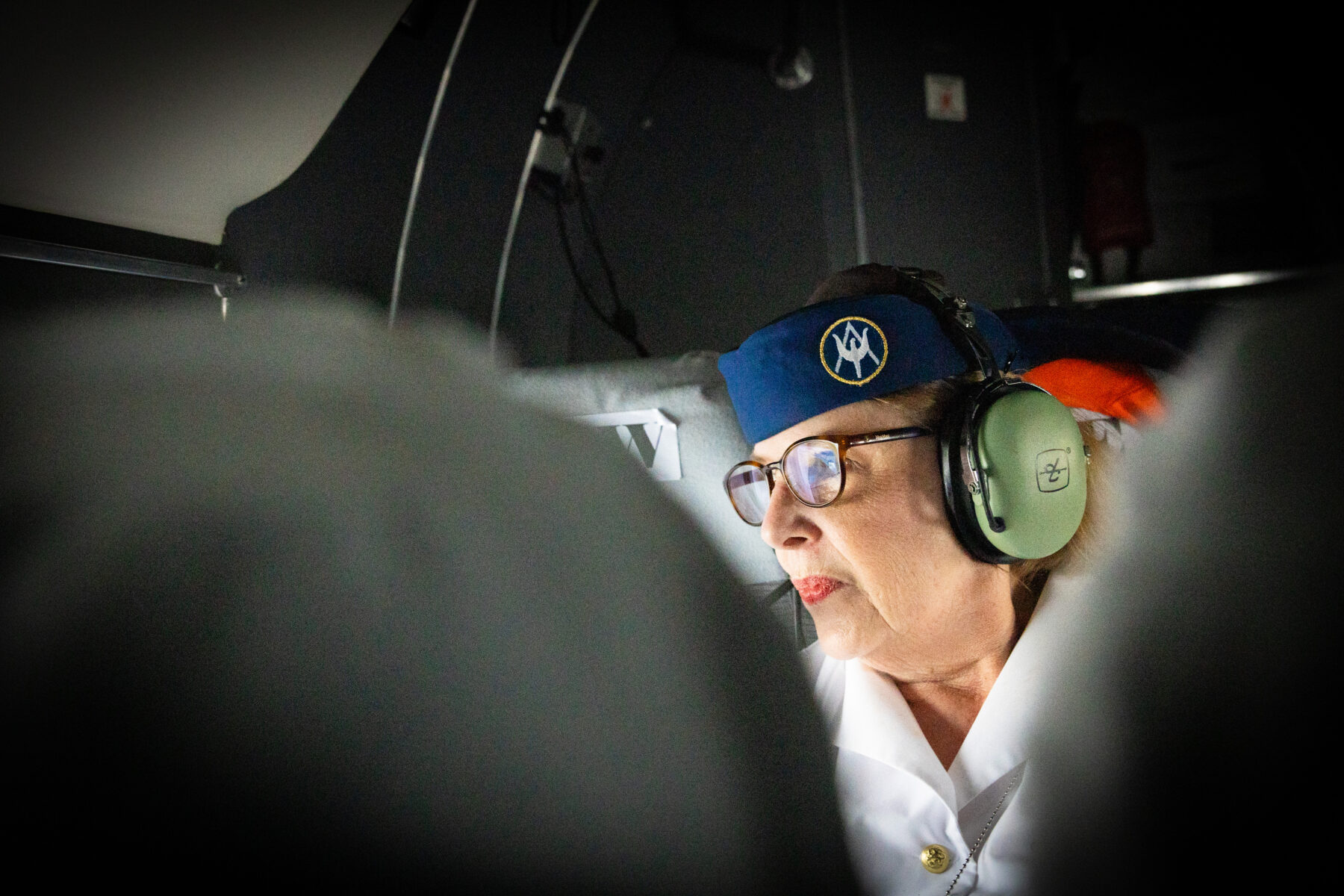 Uma comissária de bordo, vestindo um uniforme antigo e fones de ouvido, olha pela janela do avião.