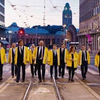 Onze adultes portant des cirés jaunes sur des costumes ou tailleurs noirs marchent en direction de l’objectif du photographe tandis que le bâtiment de la Gare principale d’Helsinki se profile derrière eux.
