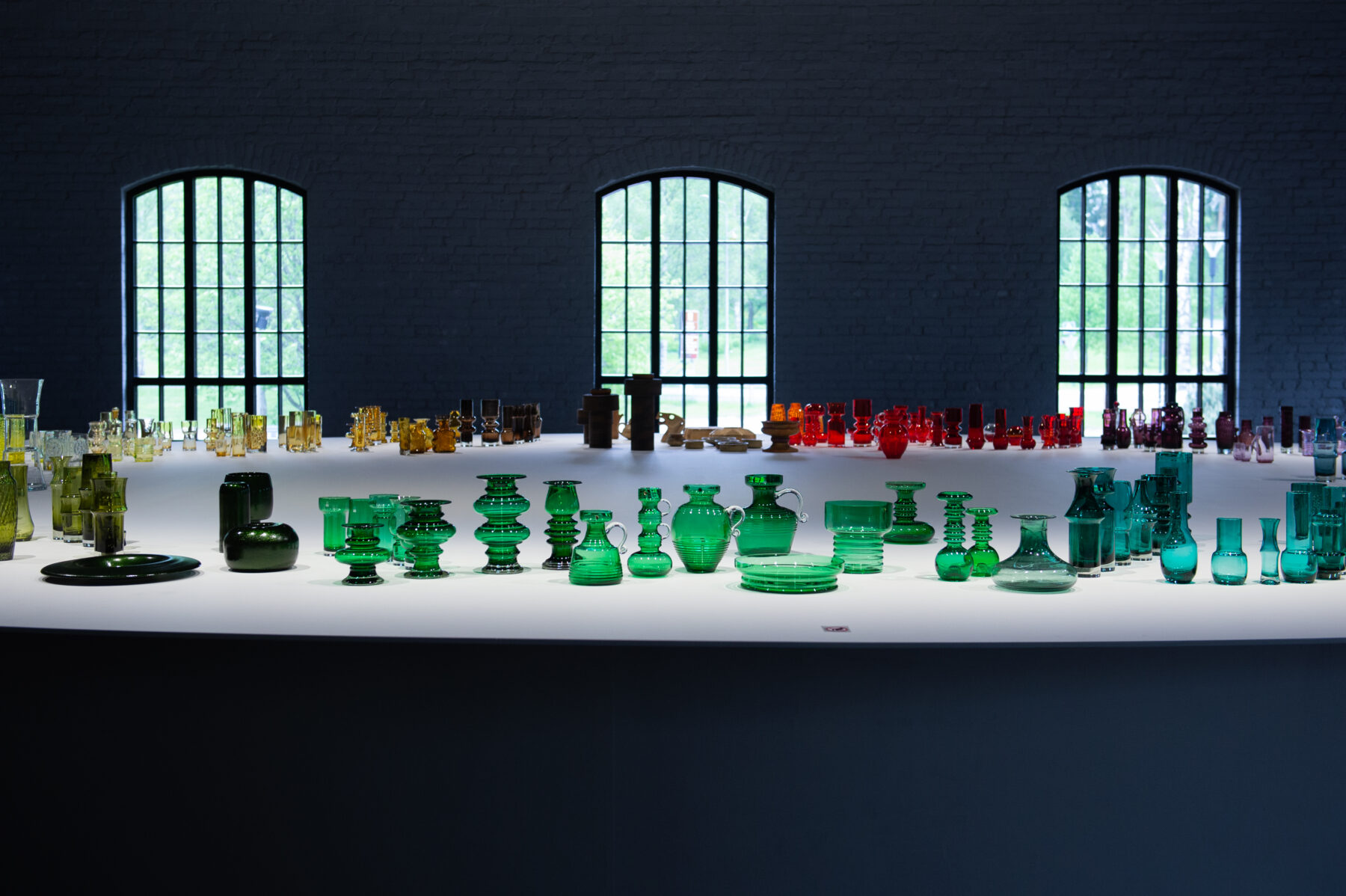 Un grand nombre de vases colorés en verre soufflé à la main sont disposés sur une longue table blanche devant trois hautes fenêtres cintrées.