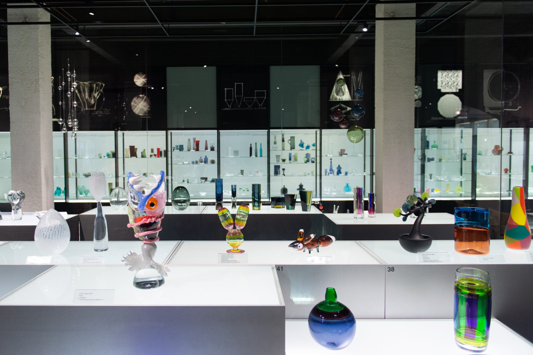 Dans une vaste salle de musée, divers objets d'art en verre sont exposés sur des tables blanches sous un éclairage vif.