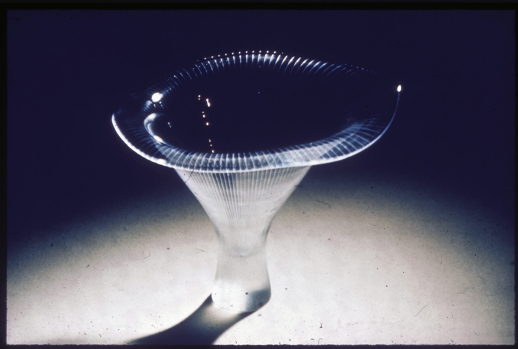 Une sculpture en verre transparent brillant dotée d'un sommet large et plat et d'une base fine striée, se détache sur un fond sombre.