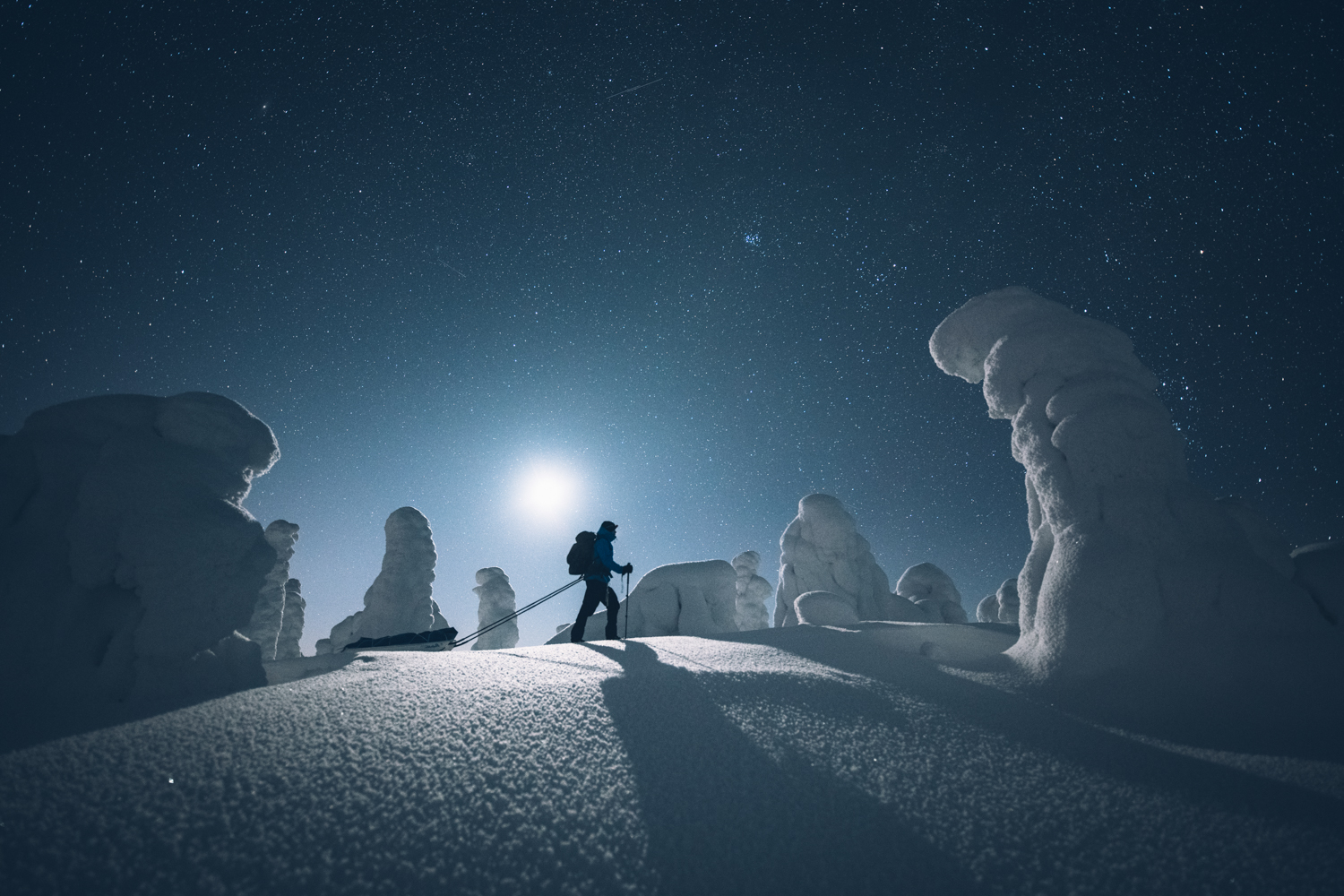 A person with a backpack and ski poles walks through a snowy landscape at night, illuminated by the moon and stars.