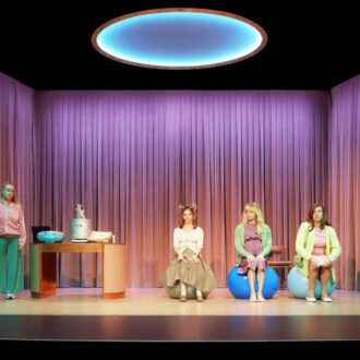 Two women and two men, all portraying women, perform on a theatre stage, three of them sitting on exercise balls and one standing near a table with objects.