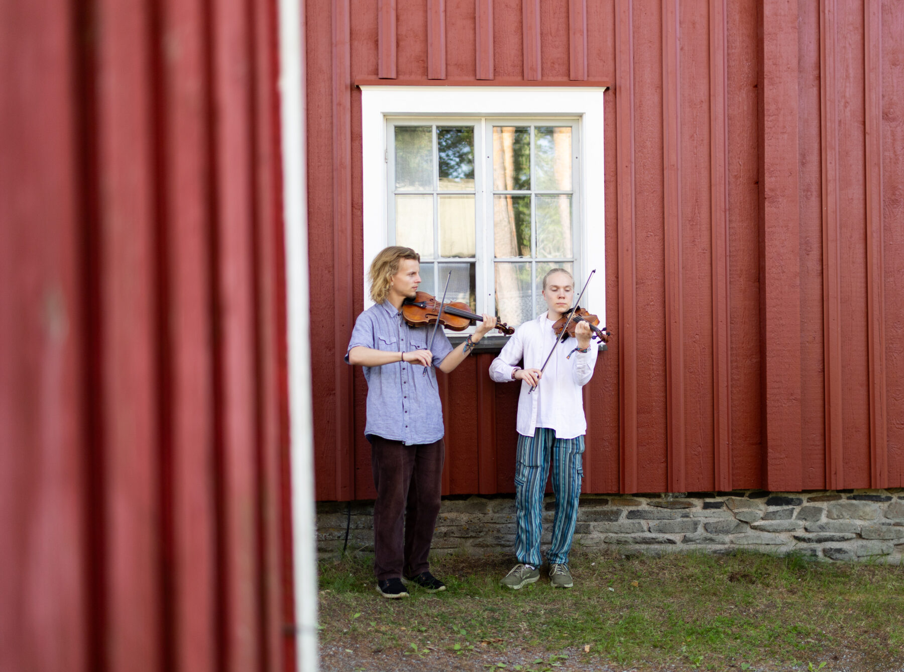 Two people play violins outside a red wooden building with a white-framed window.