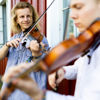 Two musicians play violins outside a red building, one in focus and the other slightly blurred in the foreground.