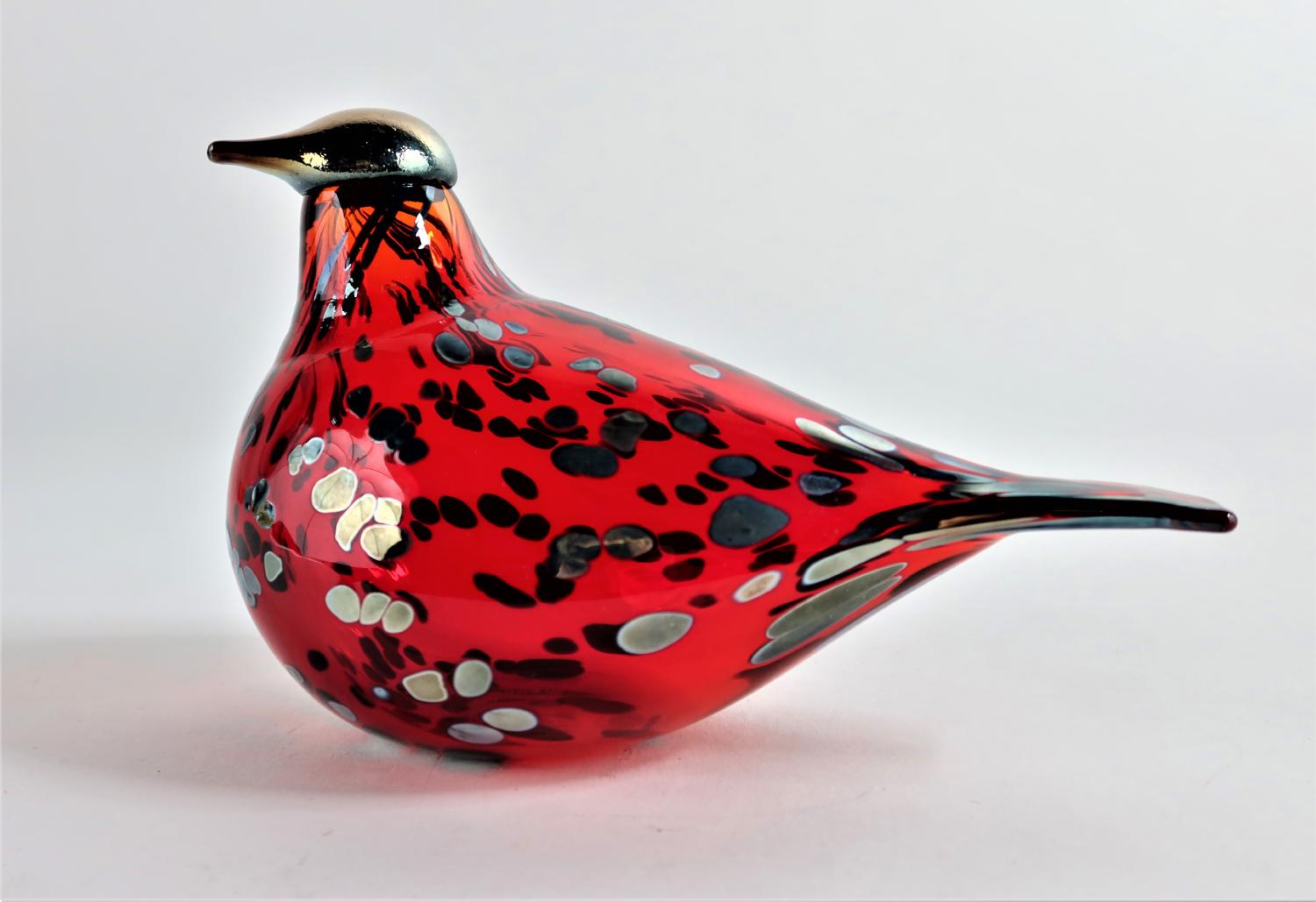 Une figurine décorative d’oiseau en verre rouge translucide mouchetée de noir se découpe sur un fond gris clair.