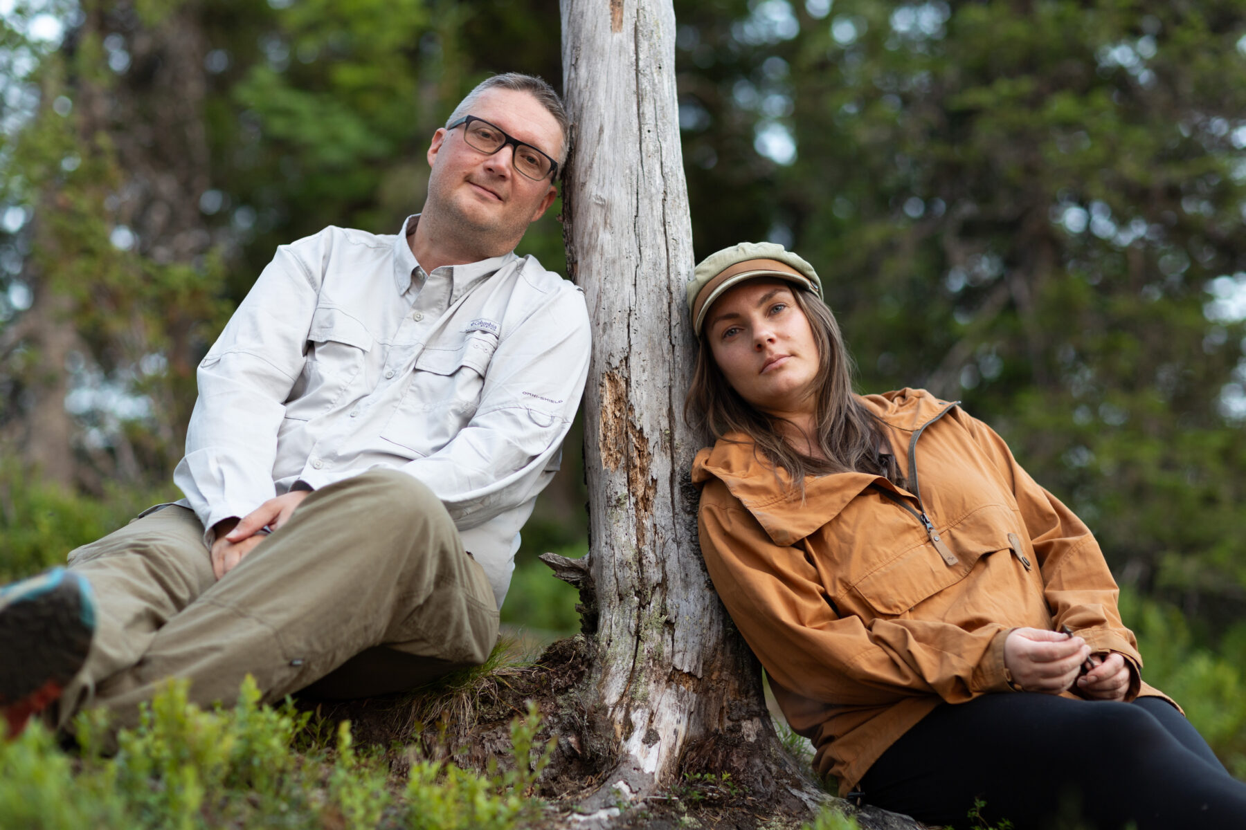 A man and a woman sit leaning against a tree trunk in a wooded area.
