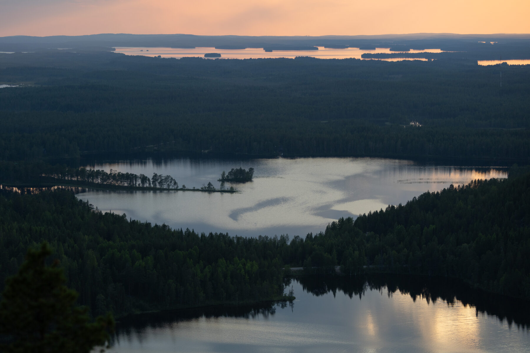 A landscape view shows a lake surrounded by a dense forest with water reflecting the sky under evening light.
