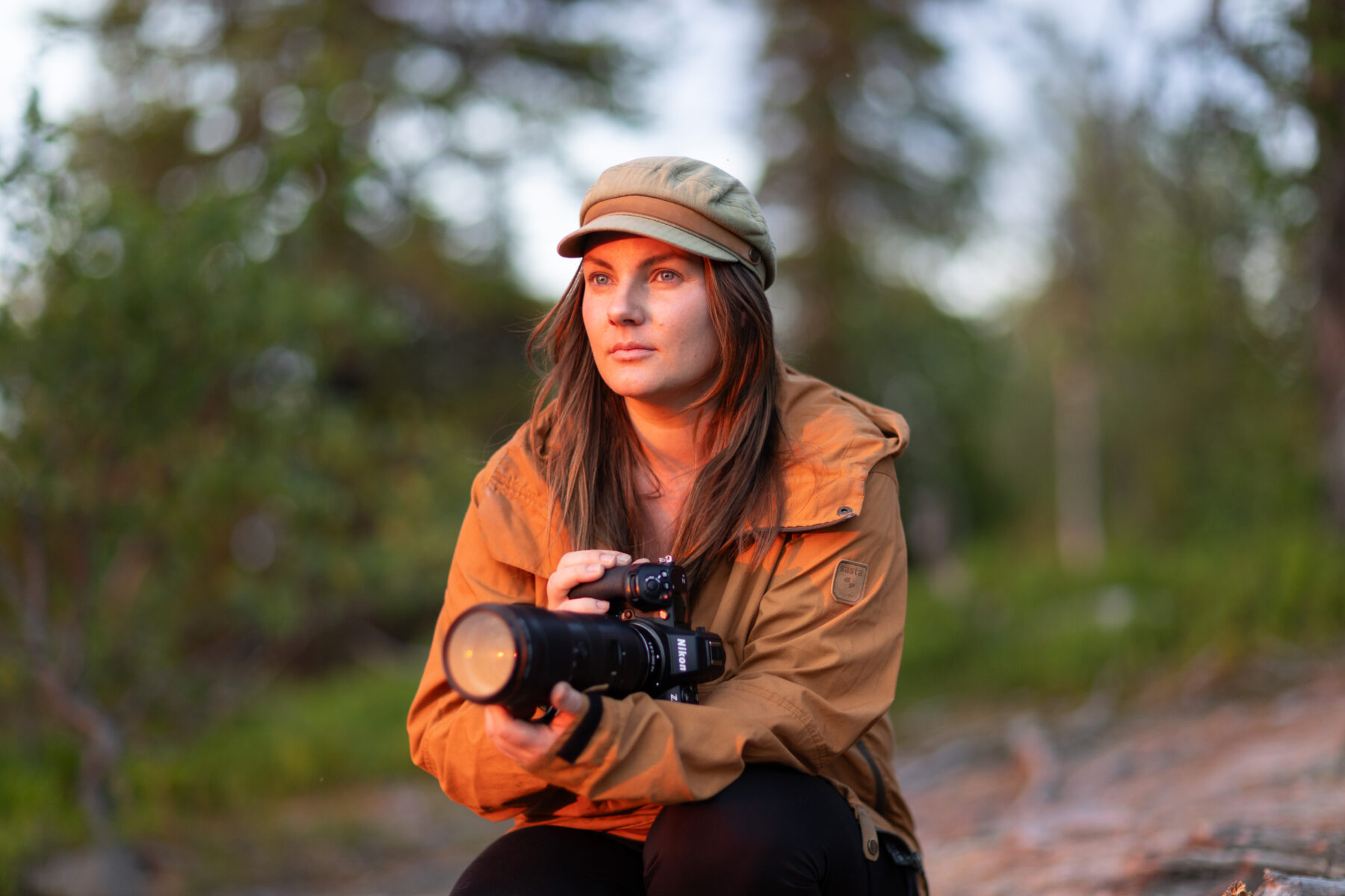 A woman wearing a jacket and hat outdoors holds a camera with a telephoto lens.