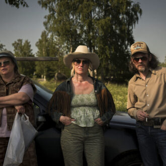 Three actors wearing hats and sunglasses lean against a car in a rural setting on a sunny day.