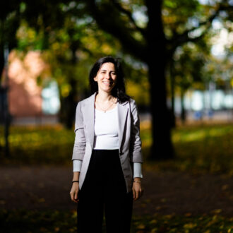 A woman standing outdoors, smiling in sunlight, with trees and green foliage in the background.