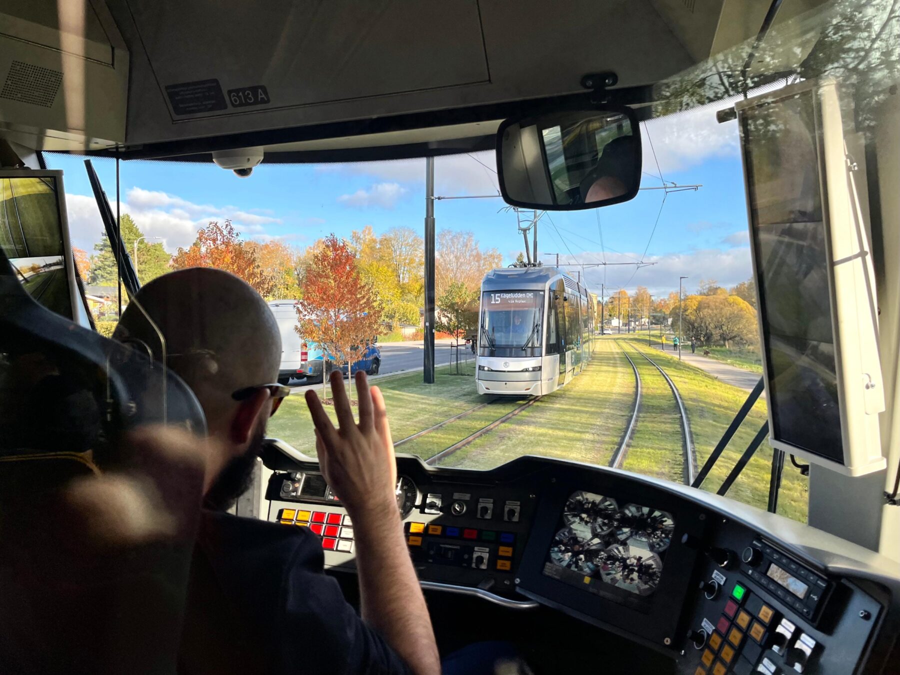 A tram operator waves from an open view of the driver's cabin with another tram car visible outside on tracks.