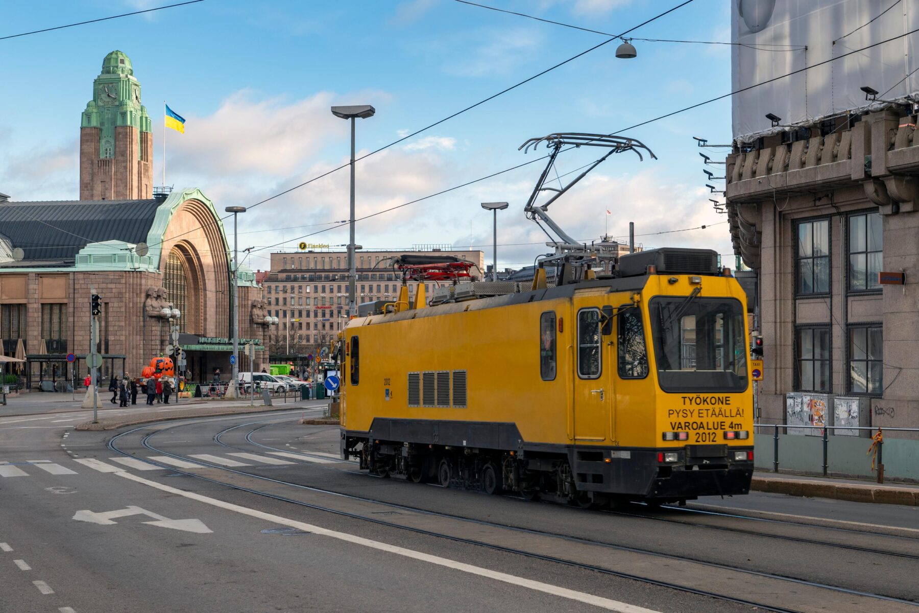 A yellow maintenance tram traverses an intersection in an urban setting, with pedestrians and grand architecture visible under a blue sky.