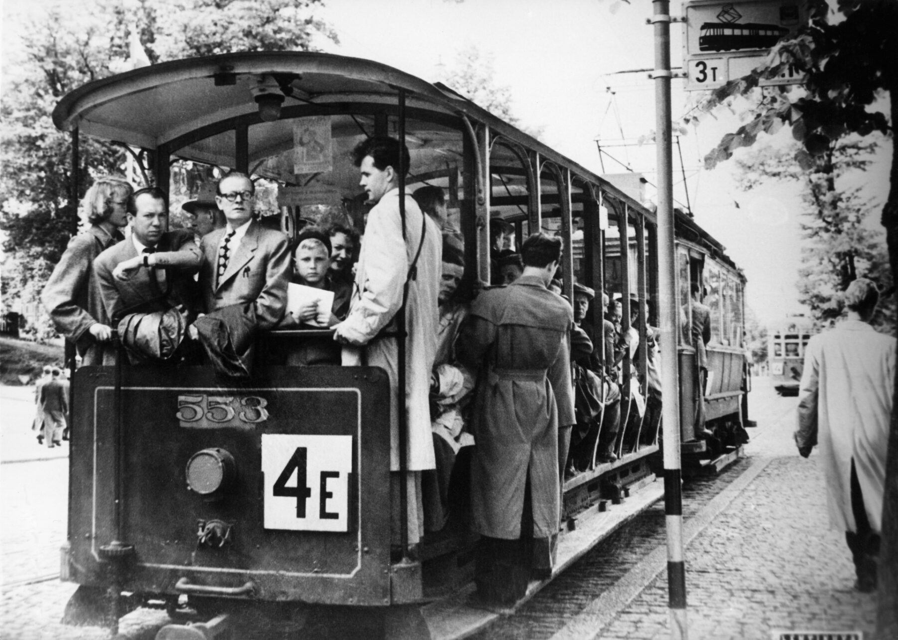 A black and white picture shows a crowd of people dressed in 1950s clothing crammed onto an open-air tram on a cobblestone city street.